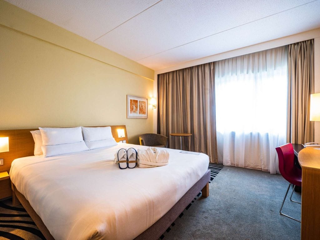 A bright hotel room with a large bed, white linens, two towel swans, a window with beige curtains, a desk with a red chair, a gray armchair, and modern wall art above the bed.