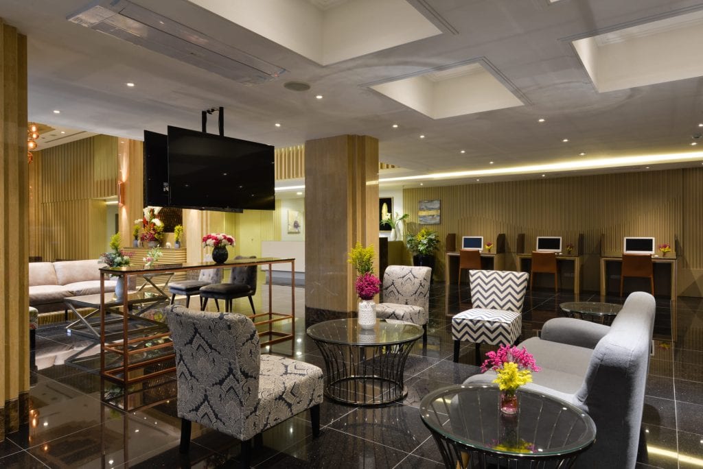 Modern hotel lobby with stylish seating, glass tables, patterned armchairs, fresh flower arrangements, and a row of computer stations in the background. Warm lighting and contemporary decor create a welcoming atmosphere.