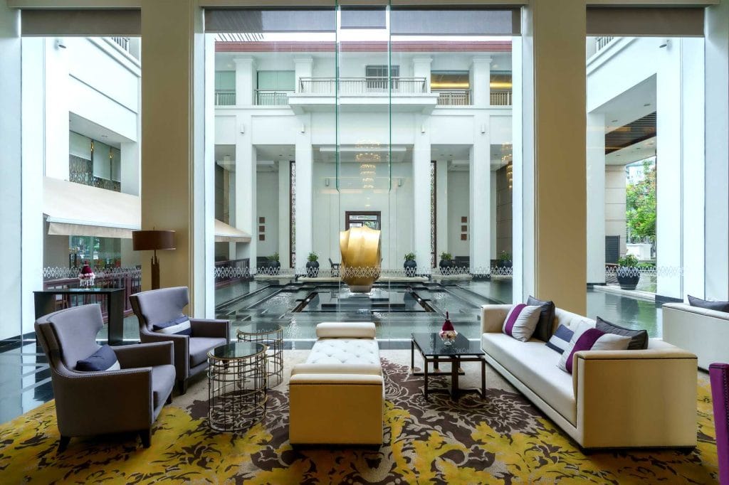 Modern hotel lobby with large glass windows, elegant seating including armchairs, a sofa, and ottomans, decorative lighting, and a view of a courtyard with a water feature and tall columns outside.