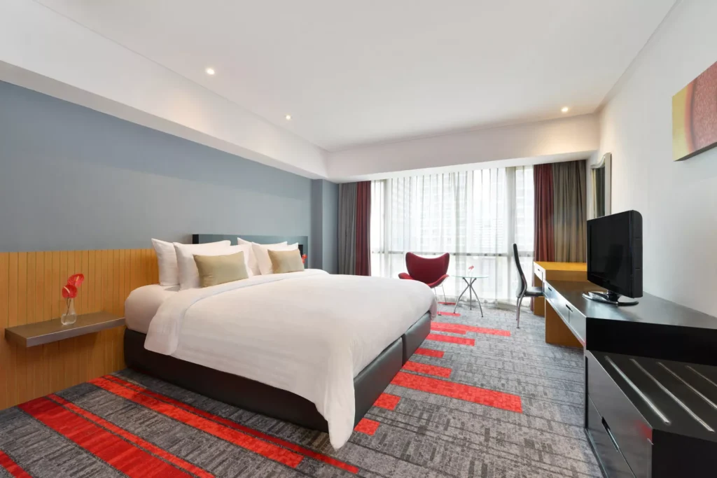 Modern hotel room with a large bed, white bedding, accent pillows, side tables with red vases, a red chair, a desk by the window, a flat-screen TV, and abstract red and gray carpet and wall art.