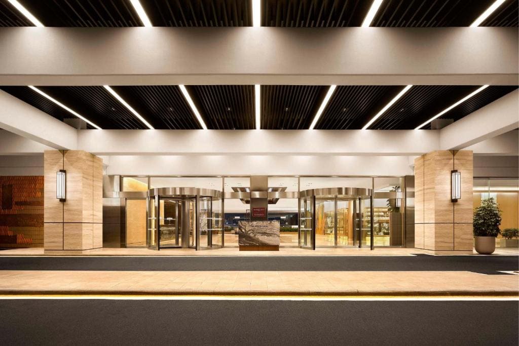 The image shows the entrance of a modern hotel with tall glass revolving doors, warm lighting, beige stone walls, and a wide driveway in front. Potted plants are placed near the entrance.
