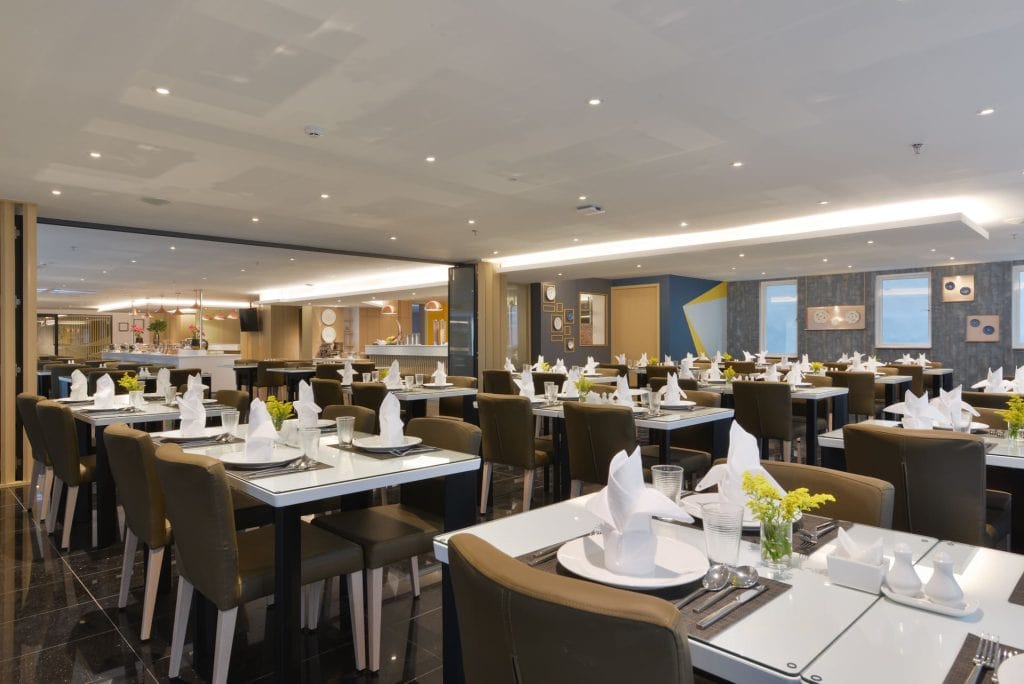 A modern, spacious restaurant dining area with neatly arranged tables. Each table is set with white napkins, plates, glasses, and small yellow flower centerpieces. The room is brightly lit with contemporary decor.