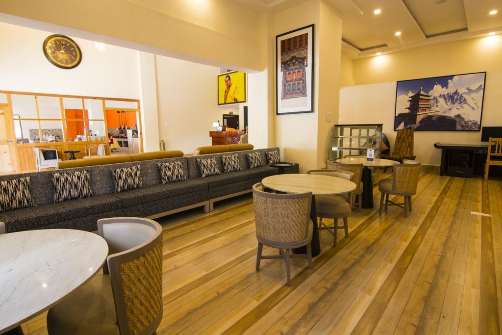 A modern lounge with wooden floors, round tables, wicker chairs, a long cushioned sofa, wall art with landscapes, and a clock above a partitioned area in the background.