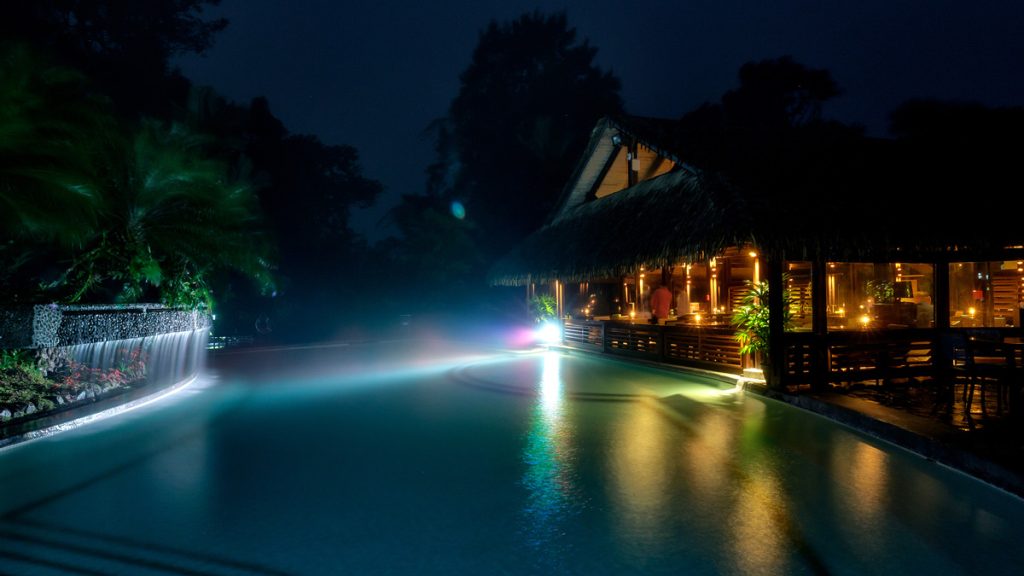A tropical pool at night with a thatched-roof restaurant beside it; warm lights glow from the building, water cascades from a small waterfall, and mist lingers above the illuminated pool.