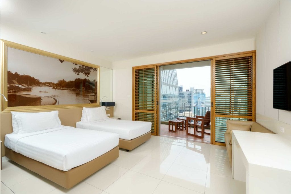 Modern hotel room with two single beds, large wall art above the headboards, sliding glass doors leading to a balcony with outdoor seating, and a cityscape view. The room is bright with white walls and tiled floor.