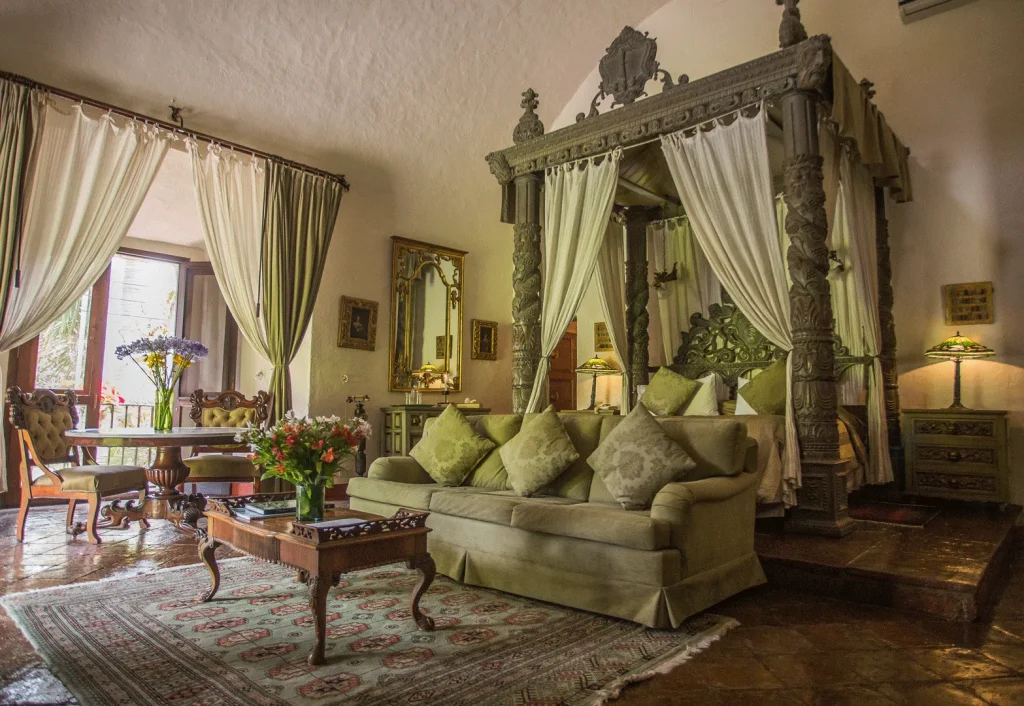 A luxurious bedroom with a carved wooden canopy bed, elegant sofa, ornate tables, fresh flowers, and tall windows with flowing curtains, creating a warm, inviting, and vintage atmosphere.