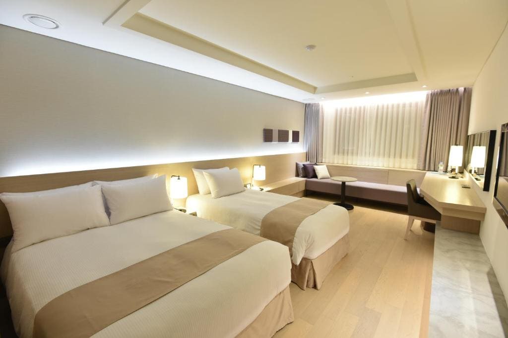 Modern hotel room with two double beds, beige bedding, a seating area by the window, a work desk with a mirror, light wood flooring, and soft, ambient lighting.