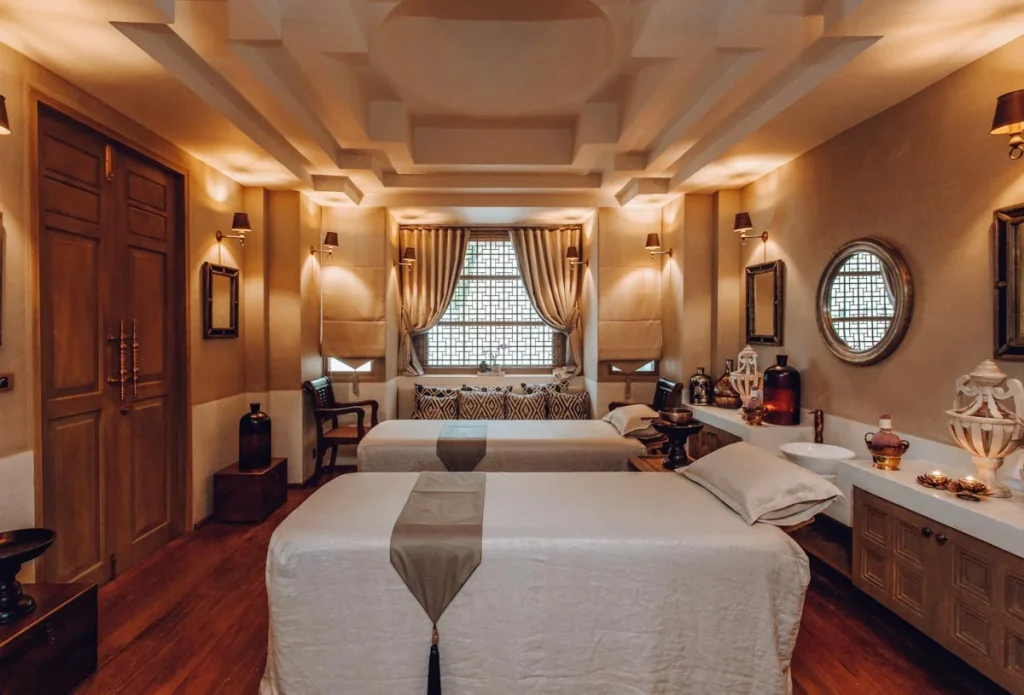 A luxurious spa room with two massage tables, soft beige linens, warm lighting, decorative vases, and a central window with curtains. Mirrors and candles add to the serene, elegant atmosphere.