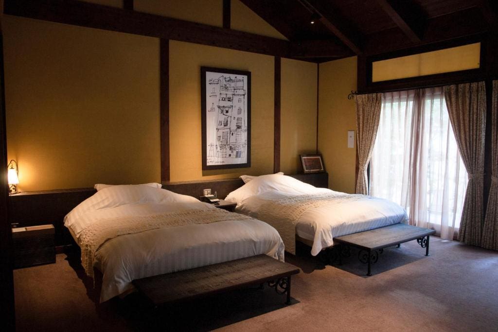 A cozy bedroom with two double beds, white bedding, wooden benches at the foot of each bed, a framed artwork above, large window with curtains, and warm yellow walls with dark wood accents.