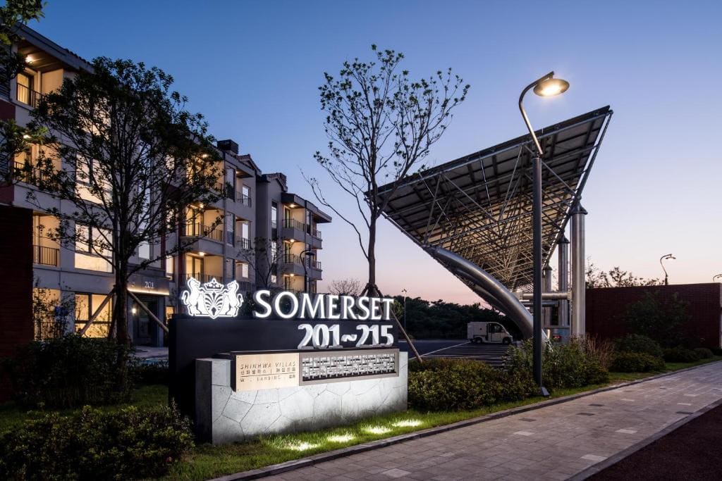 Modern apartment complex at dusk with illuminated Somerset sign, building numbers 201-215, trees, and a large metal canopy structure near the entrance. The sky is clear with streetlights on.