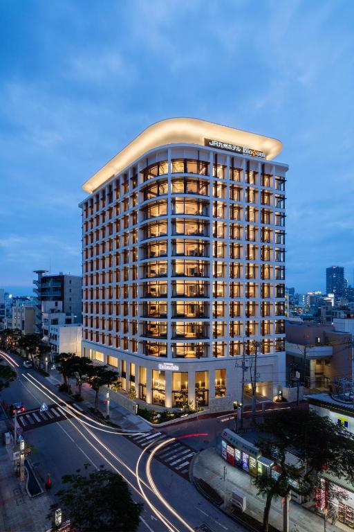 A modern, multi-story hotel with illuminated windows stands at a street corner at dusk. Light trails from moving vehicles curve around the building, and city structures are visible in the background under a cloudy sky.