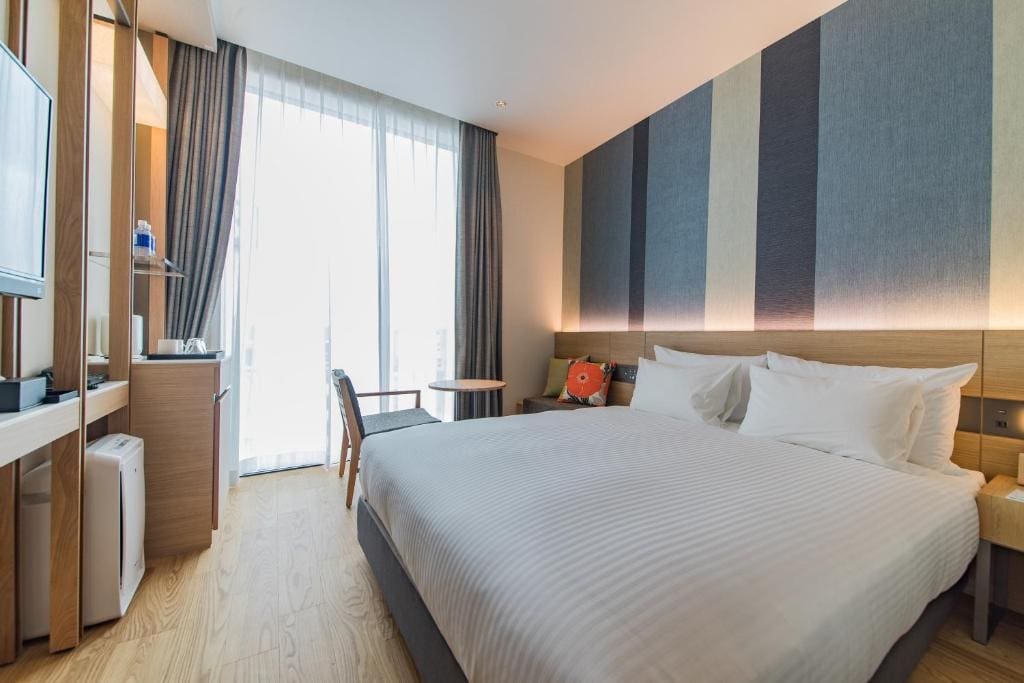 Modern hotel room with a large bed, striped accent wall, wooden floor, small desk, chair, TV, and floor-to-ceiling window with sheer curtains letting in natural light.
