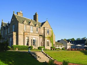 A large stone manor with tall chimneys and many windows sits on a well-manicured lawn with steps leading up to it. Outdoor seating and tables are visible to the side under a clear blue sky.