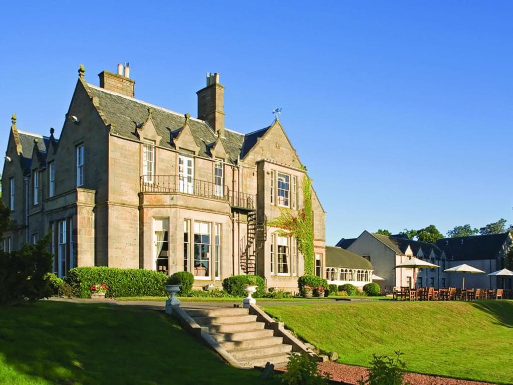 A large stone manor with tall chimneys and many windows sits on a well-manicured lawn with steps leading up to it. Outdoor seating and tables are visible to the side under a clear blue sky.