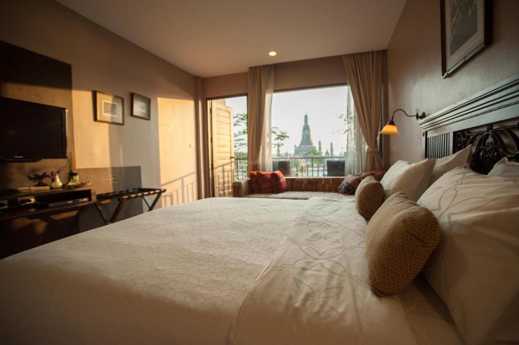 A cozy hotel room with a large bed, soft pillows, TV, and window seating. The window offers a scenic view of a landmark temple in the distance, illuminated by warm natural light.