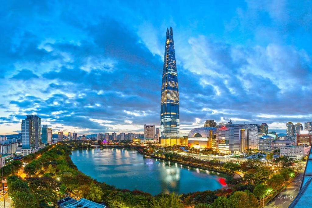 A tall skyscraper rises above a city skyline at dusk, surrounded by modern buildings, bright city lights, and a large lake with trees lining the shore under a dramatic, cloudy blue sky.