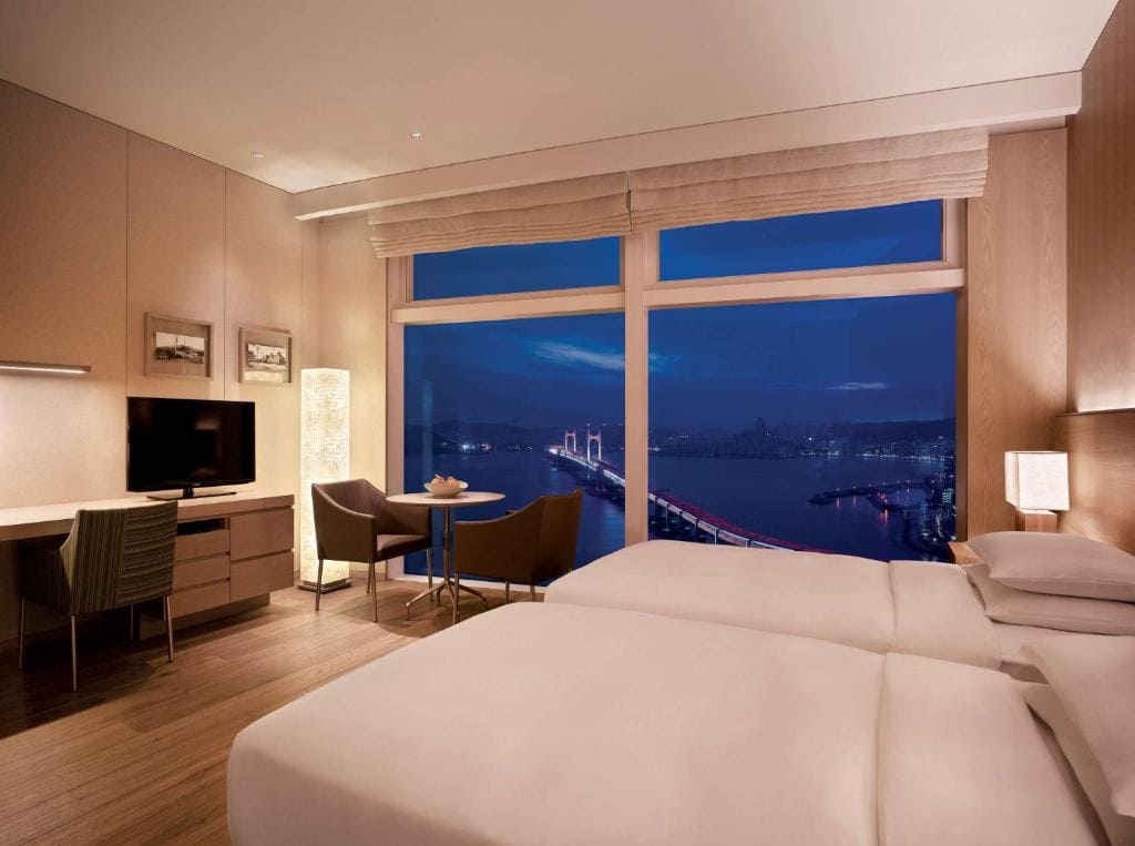 A modern hotel room with two beds, a desk, a TV, and a small dining area by a large window overlooking a cityscape and a lit-up bridge at dusk. The room features warm lighting and minimalist decor.