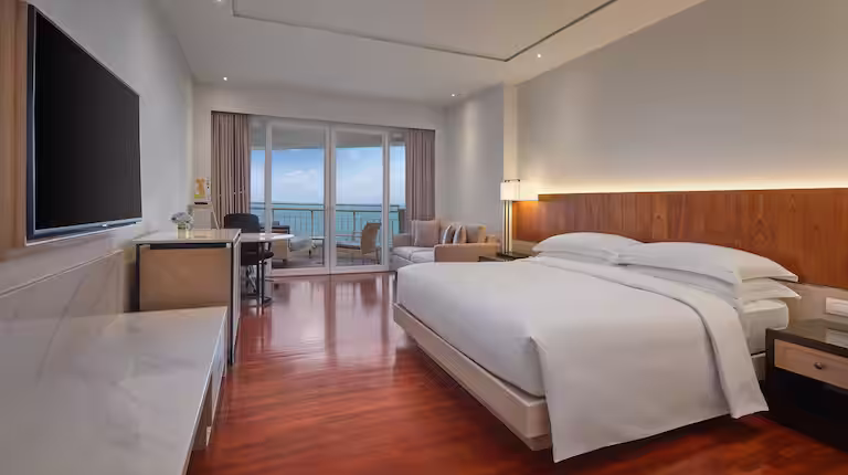 A modern hotel room with a large bed, wooden floors, a flat-screen TV, desk, seating area, and glass doors leading to a balcony with an ocean view.