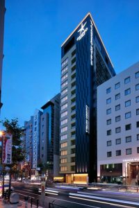 A tall, modern hotel building with a sleek black exterior and illuminated signs stands among other city buildings at dusk, with blurred car lights on the street below.