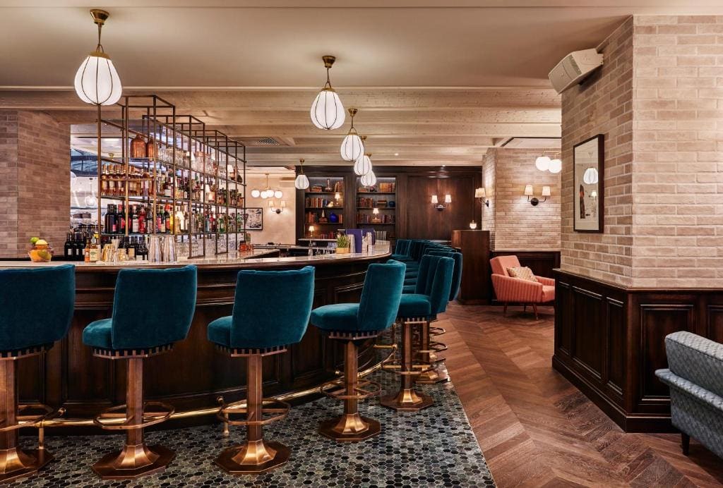 A stylish bar with teal velvet stools, a dark wood counter, and hanging pendant lights. Shelves of bottles are behind the bar, and there is a cozy lounge area with armchairs in the background.