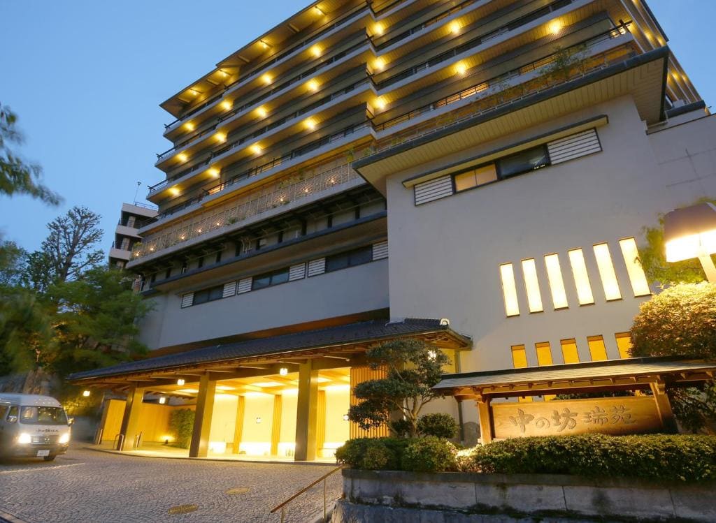 A large, multi-story hotel building is brightly lit in the evening, with warm yellow lights illuminating the entrance and balconies. A van is parked outside on the cobblestone driveway surrounded by greenery.