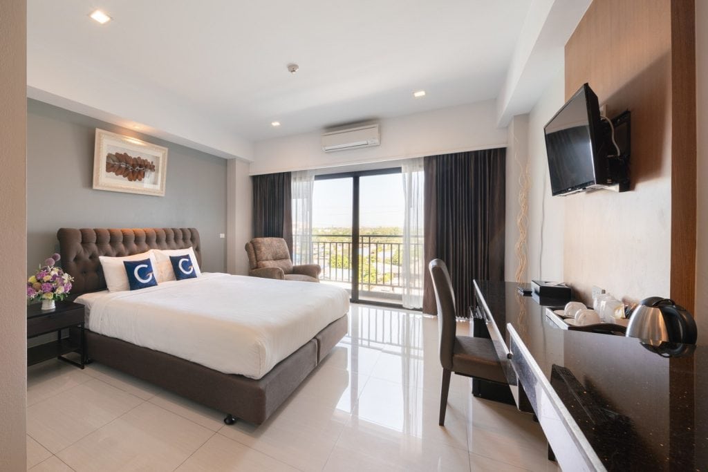 Modern hotel room with a large bed, two pillows, side tables, an armchair, a wall-mounted TV, a desk, tea and coffee facilities, and a balcony with sunlight streaming through glass doors.