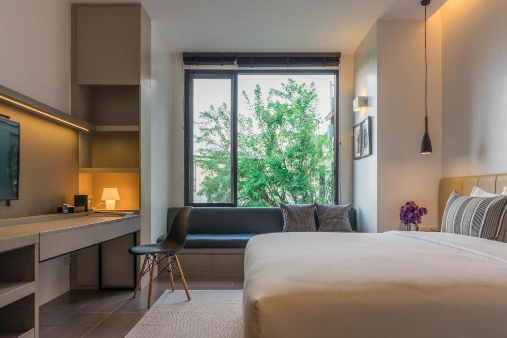 Modern hotel room with a large bed, a black chair, a desk with a lamp, a wall-mounted TV, and a window seat beneath a large window showing green trees outside. Minimal decor, soft lighting, and neutral colors.