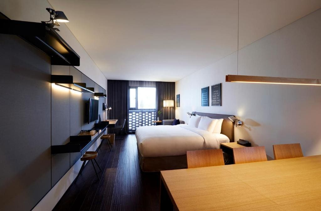 A modern hotel room with a large bed, a desk, a wall-mounted TV, wooden flooring, and a window with dark curtains letting in natural light. Minimalistic decor and warm lighting create a cozy atmosphere.
