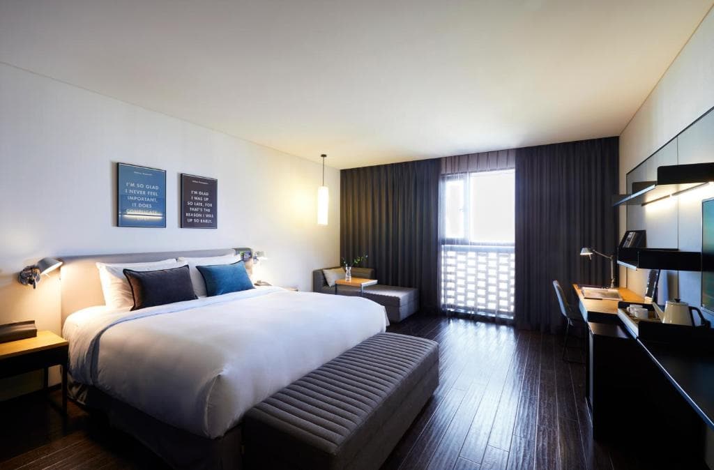 Modern hotel room with a large bed, blue and white bedding, two framed pictures above the headboard, a padded bench at the foot of the bed, a desk with a chair, and a window with dark curtains letting in light.