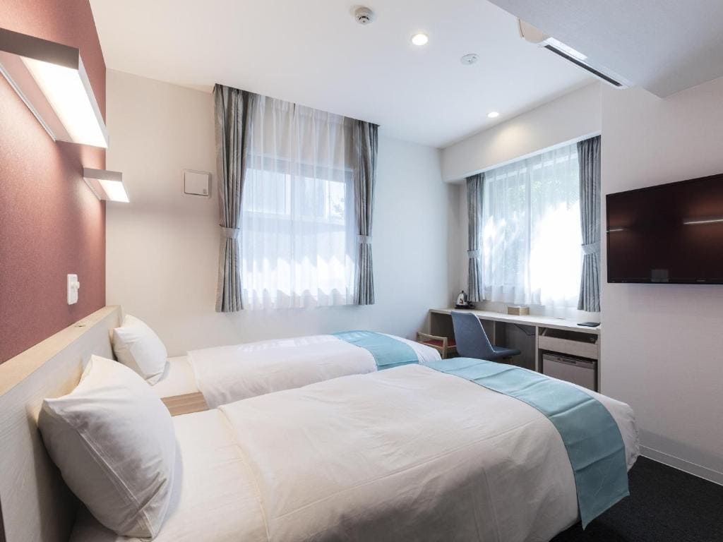 A modern hotel room with two single beds, white bedding with light blue runners, two large windows with curtains, a wall-mounted TV, a desk, and minimalistic decor.