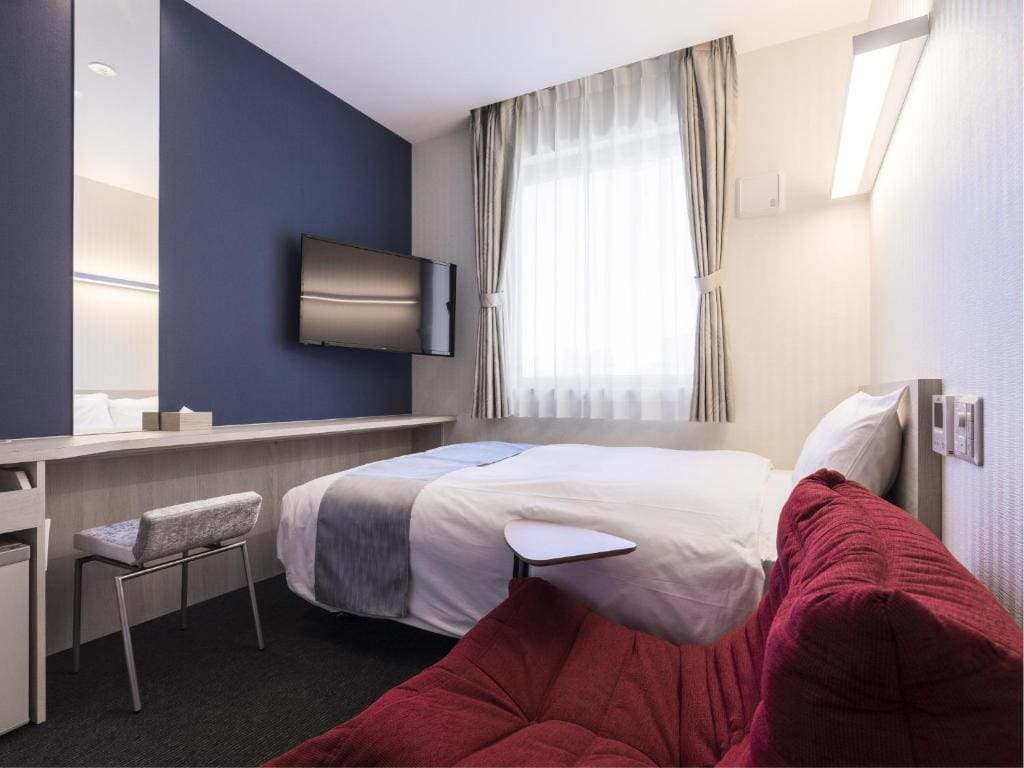 Modern hotel room with a double bed, wall-mounted TV, large mirror above a desk with a chair, a window with curtains, and a red cushioned sofa in the foreground. The decor is minimalist and tidy.