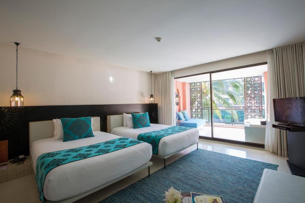 A modern hotel room with two twin beds, turquoise accents, a large window with sheer curtains, a flat-screen TV, and a view of palm trees and a balcony outside.