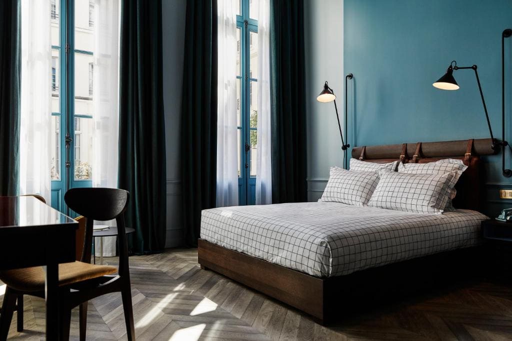 A modern bedroom with tall windows, teal curtains, a blue accent wall, and a double bed with checkered bedding. Black wall lamps, a desk, and a chair are also visible. Sunlight streams through the sheer curtains.