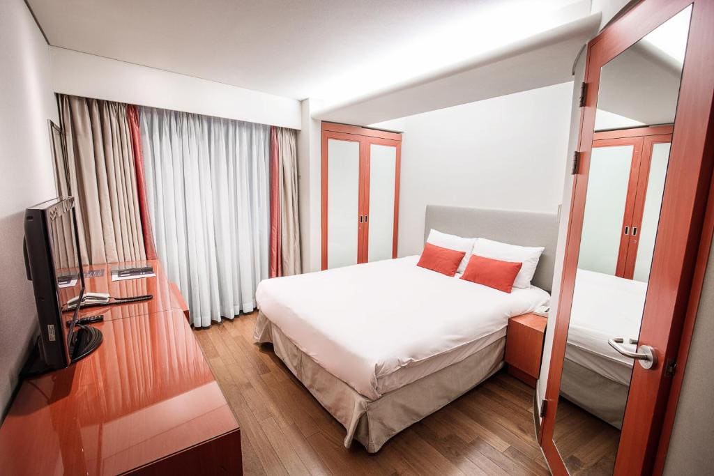 A modern hotel room with a double bed, white bedding, and red accent pillows. The room features wooden floors, red furniture, a flat-screen TV on a desk, and large closets with mirrored doors. Curtains cover the window.