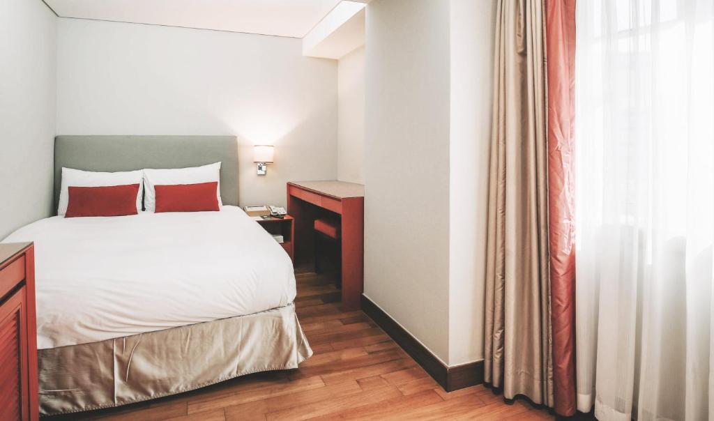 A bright hotel room with a neatly made double bed, accented with red pillows, a small wooden desk and chair, and a large window covered by beige curtains letting in natural light.