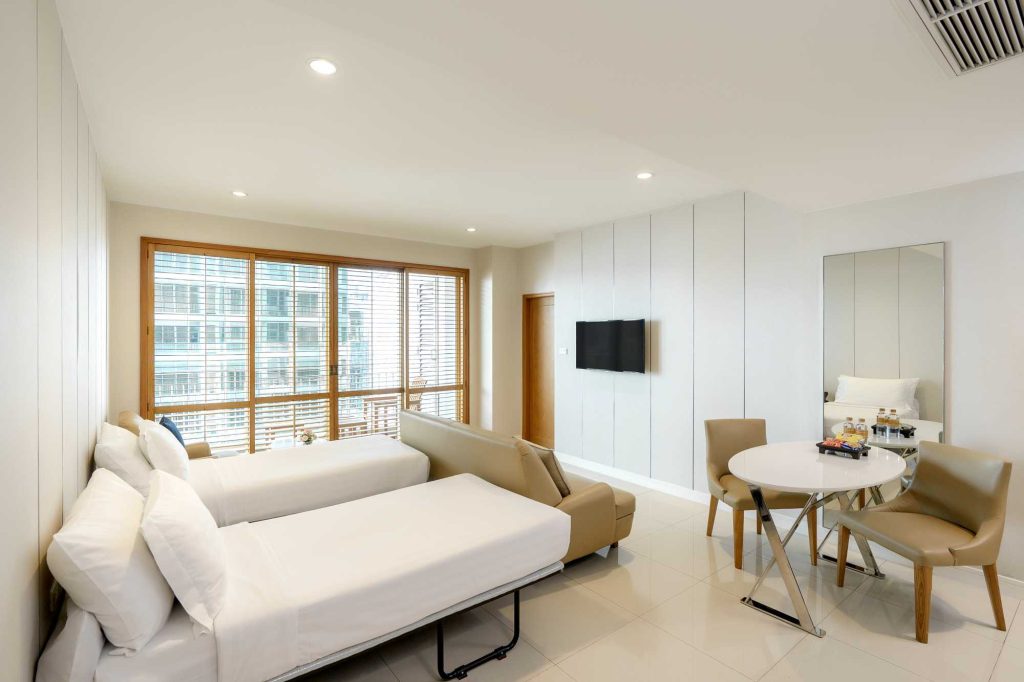 A modern hotel room with two single beds, a beige sofa, a wall-mounted TV, a round dining table with four chairs, a large mirror, and floor-to-ceiling windows with blinds letting in natural light.