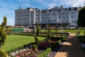 A grand grey hotel labeled The Hythe Imperial stands behind a landscaped garden with green lawns, colorful flowerbeds, small trees, and fountains under a clear blue sky.