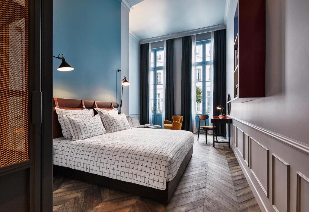 Modern bedroom with tall windows, blue walls, a large bed with checkered bedding, wall-mounted lamps, a yellow armchair, a desk with a chair, and herringbone wood flooring. Natural light fills the room.