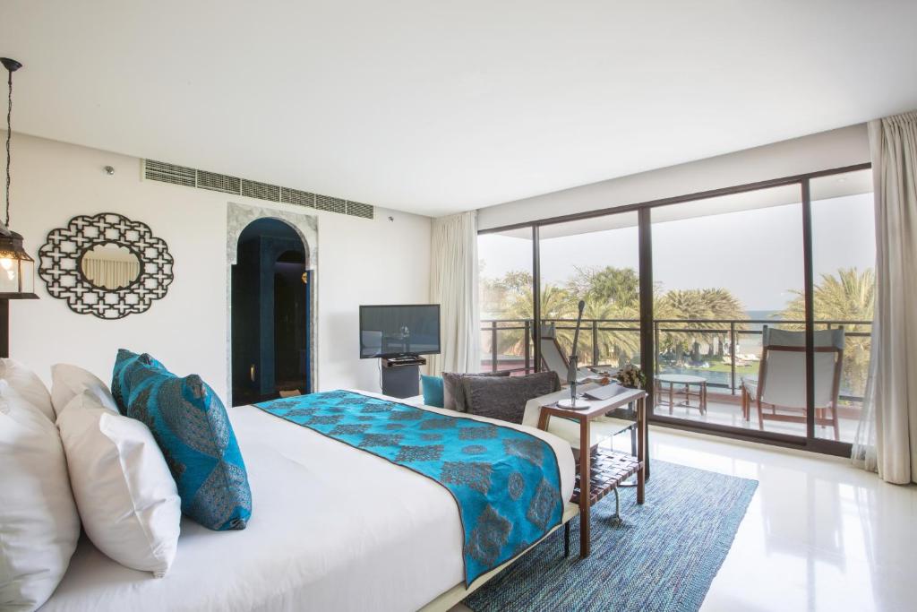 A modern hotel room with a large bed featuring blue and white bedding, a flat-screen TV, armchair, and a balcony with outdoor seating overlooking palm trees and a pool area. Natural light fills the room.