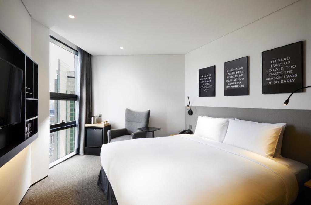 A modern hotel room with a large bed, gray chair, small fridge, wall-mounted TV, and three framed quotes above the headboard. Large windows let in natural light. The decor is minimal and stylish.