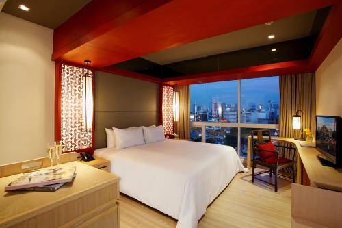 Modern hotel room with a large bed, wooden furniture, a desk, a TV, and large windows overlooking a cityscape at dusk. Warm lighting and red decorative accents create a cozy, inviting atmosphere.