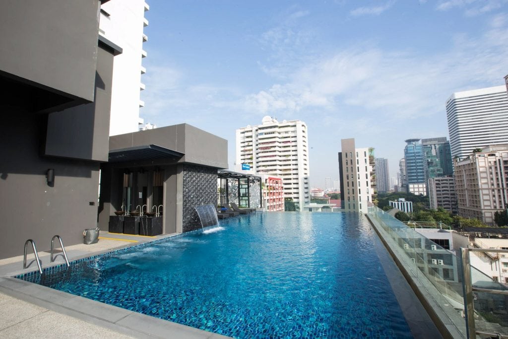 Rooftop infinity pool with clear blue water, surrounded by modern buildings and skyscrapers under a bright sky. A small waterfall feature pours into the pool near a shaded seating area.