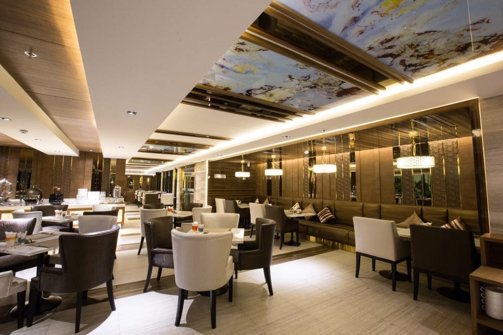 A modern, upscale restaurant interior with elegant brown and cream chairs, neatly set tables, mirrored walls, ceiling art panels, and warm ambient lighting.