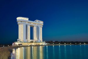 A modern, uniquely shaped high-rise hotel with two curved towers connected at the top, brightly lit at night, stands beside a calm body of water under a clear, dark blue sky.