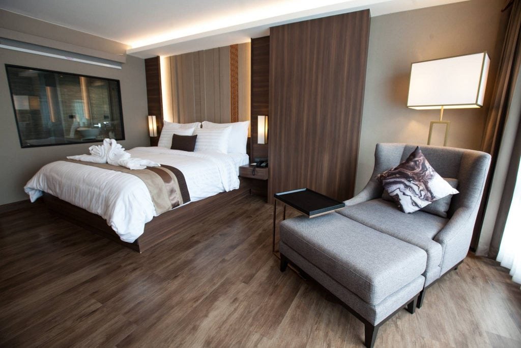 Modern hotel room with a large bed, white linens, and decorative towels. There is a gray upholstered chair with a pillow and side table, wood floors, and soft lighting, creating a cozy and inviting atmosphere.