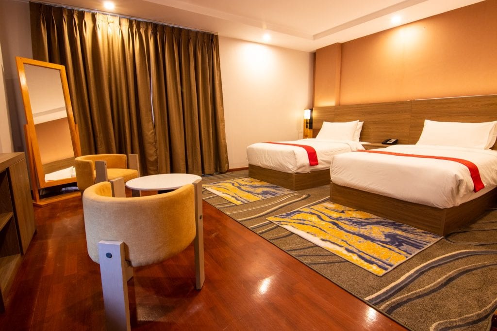 A modern hotel room with two beds, white bedding with red accents, a small table and two chairs, a standing mirror, brown curtains, and warm lighting on wood floors and patterned rugs.