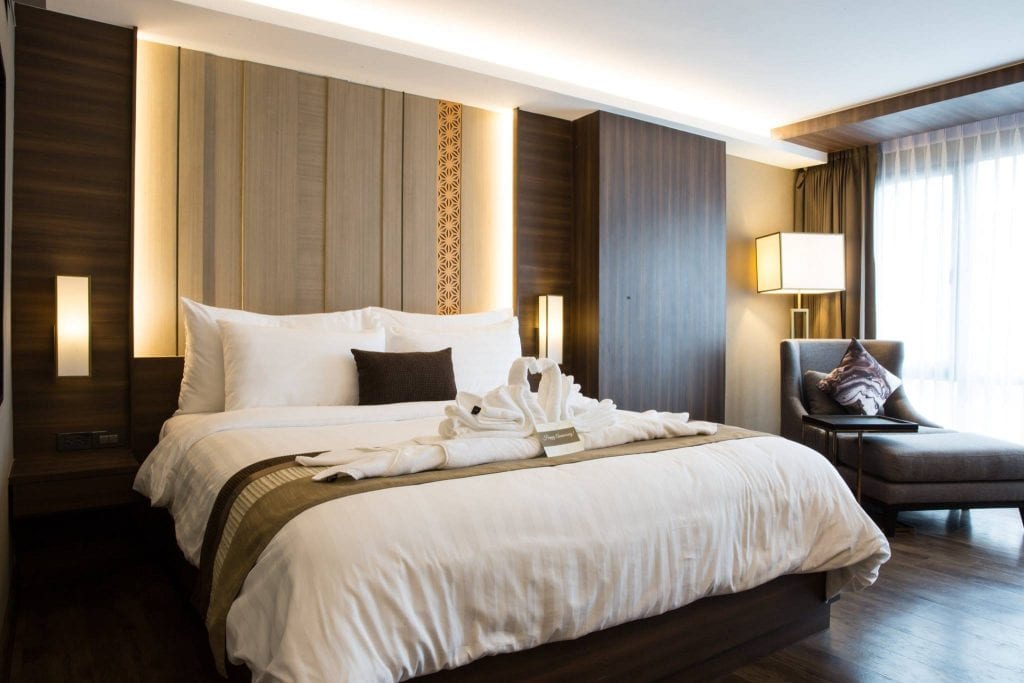 A modern hotel room with a neatly made double bed, decorative pillows, towel swans on the bed, warm lighting, wood paneling, and a window with sheer curtains beside a cushioned chair and floor lamp.