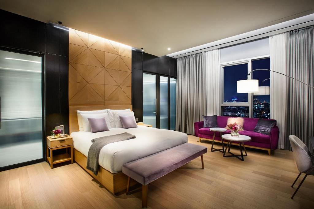 Modern hotel room with a large bed, purple bench, and pillows, a stylish purple sofa, round coffee table, and large windows showcasing a city skyline at night. Room features wood flooring and contemporary decor.