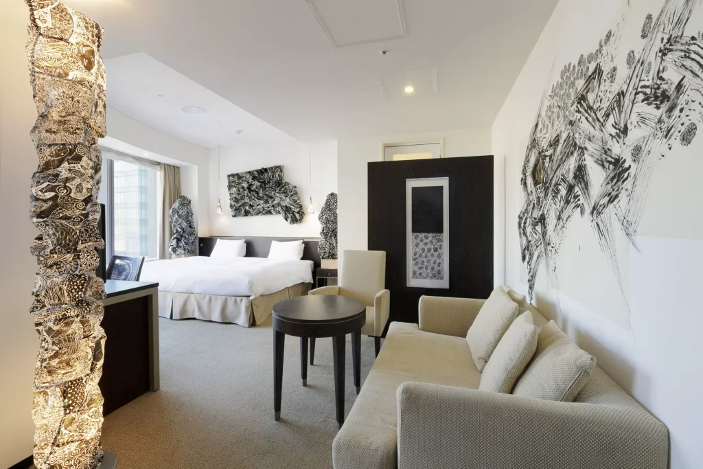 Modern hotel room with a double bed, beige sofa, round table, and unique black-and-white abstract art on the walls and lamp. Large window lets in natural light, creating a bright and spacious atmosphere.