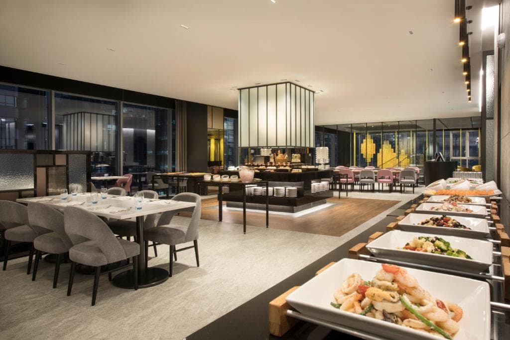 Modern, upscale buffet restaurant with elegant seating, stylish lighting, and a variety of dishes displayed in white trays on a counter; large windows offer a view of the city outside.
